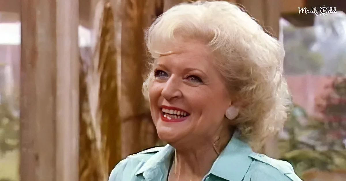 A smiling woman with blonde hair, wearing a light blue shirt, stands in a room with wooden elements and a blurred background.