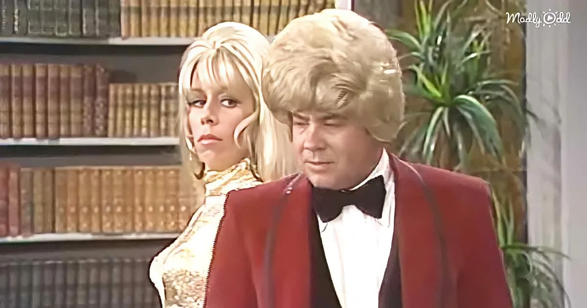 A man in a red suit and bow tie stands beside a woman in a gold dress, both in a room with bookshelves and a plant.