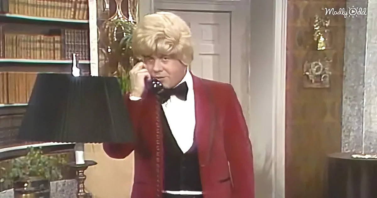 A man in a red tuxedo and blonde wig talks on a rotary phone in a vintage interior setting.