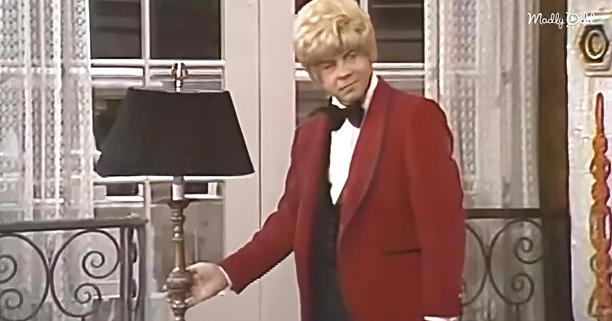 A person in a red tuxedo and white gloves stands near a lamp, with a window and decorative railing in the background.