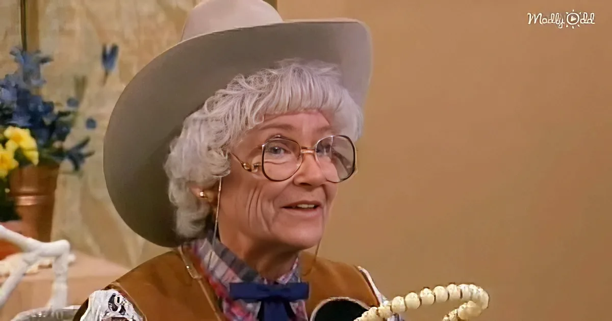 An elderly woman wearing a cowboy hat and plaid shirt holds a guitar, smiling.