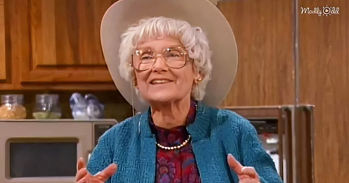 An older woman with white hair, glasses, and a blue sweater stands in a kitchen, smiling and gesturing.