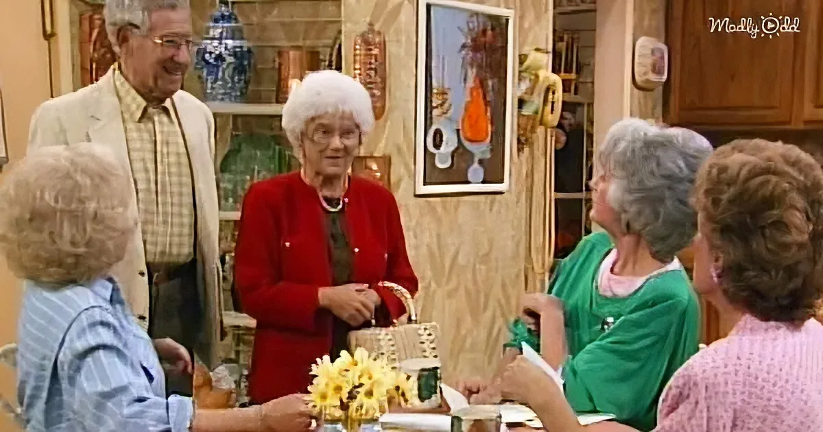 A woman in a red jacket stands in a kitchen, surrounded by other women, with a vase of yellow flowers on the table.
