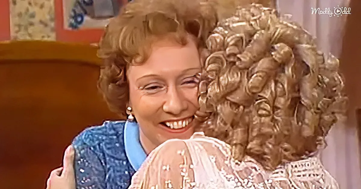 A smiling woman hugs another woman with voluminous, curly hair in a cozy indoor setting.
