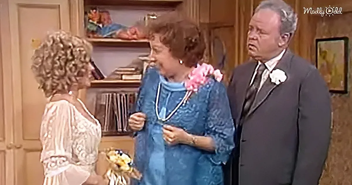 Woman in blue dress adjusts necklace, flanked by man in suit and woman in white lace dress, in a cozy room.