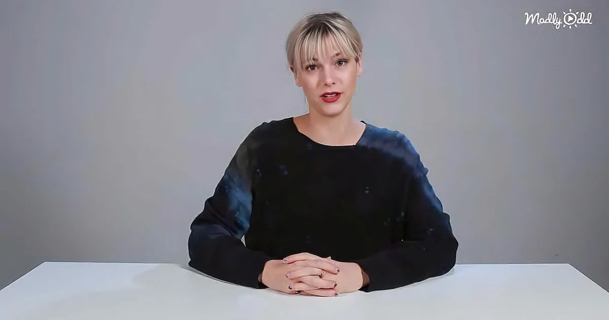 A person with short blonde hair sits at a white table, wearing a dark, speckled sweater, with hands clasped.