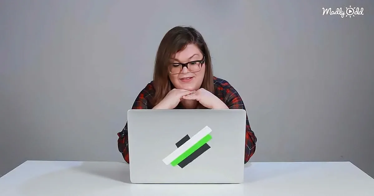 A person wearing glasses sits at a white table with a laptop, hands on chin, in front of a gray background.