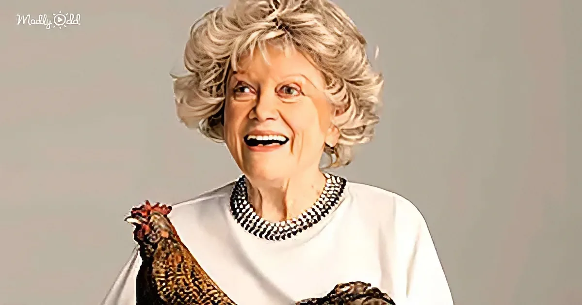 A smiling woman with short, curly blonde hair and a pearl necklace, wearing a white top, with a chicken perched on her...