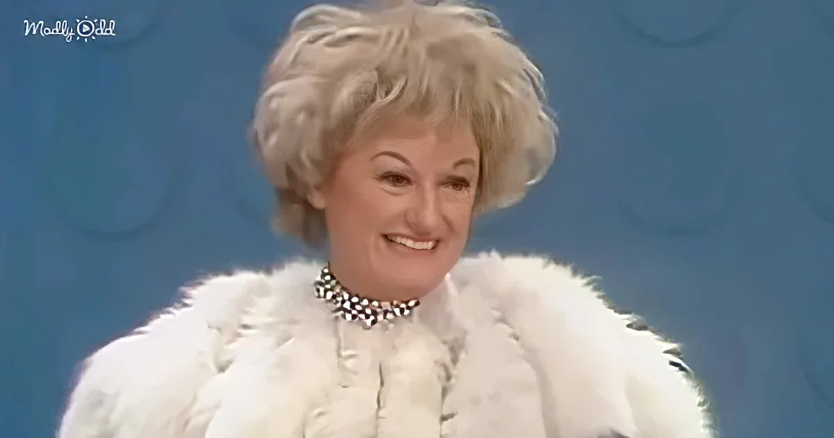 A woman with short, styled blonde hair smiles in a white fur coat and pearl necklace against a blue background.