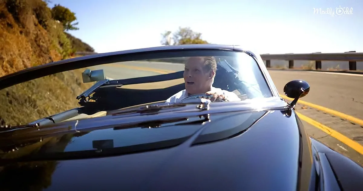 A man drives a sleek black convertible along a scenic road with a clear sky and greenery in the background.