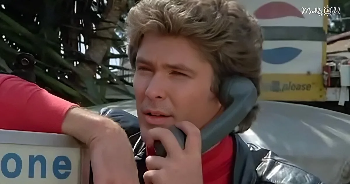 A man in a shiny jacket and headset is talking on a payphone, with palm trees and a gas station in the background.