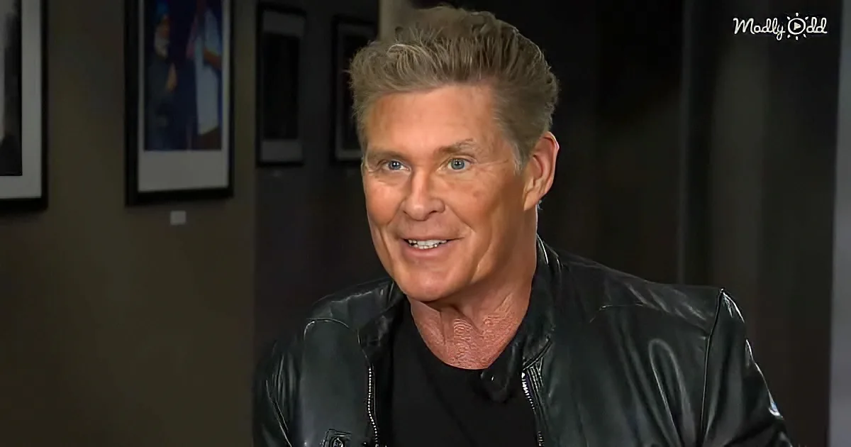 A man with short, styled hair, wearing a black leather jacket, smiles while seated in a room with framed pictures on the...