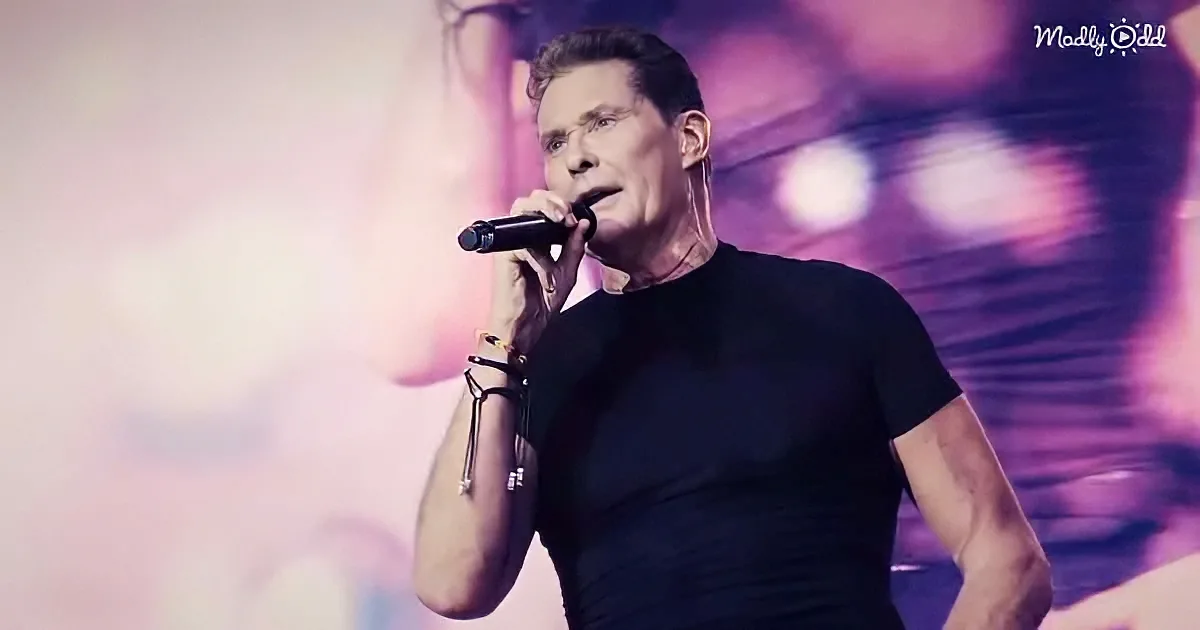 A person in a black t-shirt sings into a microphone on stage with a blurred background.