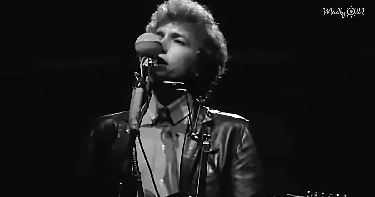 A person in a leather jacket sings into a microphone on stage.