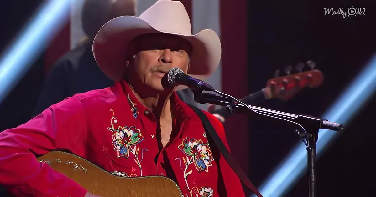 A man in a red floral shirt and cowboy hat plays guitar and sings into a microphone.