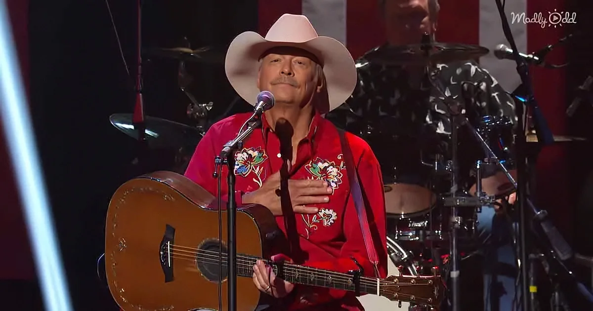 A man in a red shirt and cowboy hat plays guitar on stage, singing into a microphone.