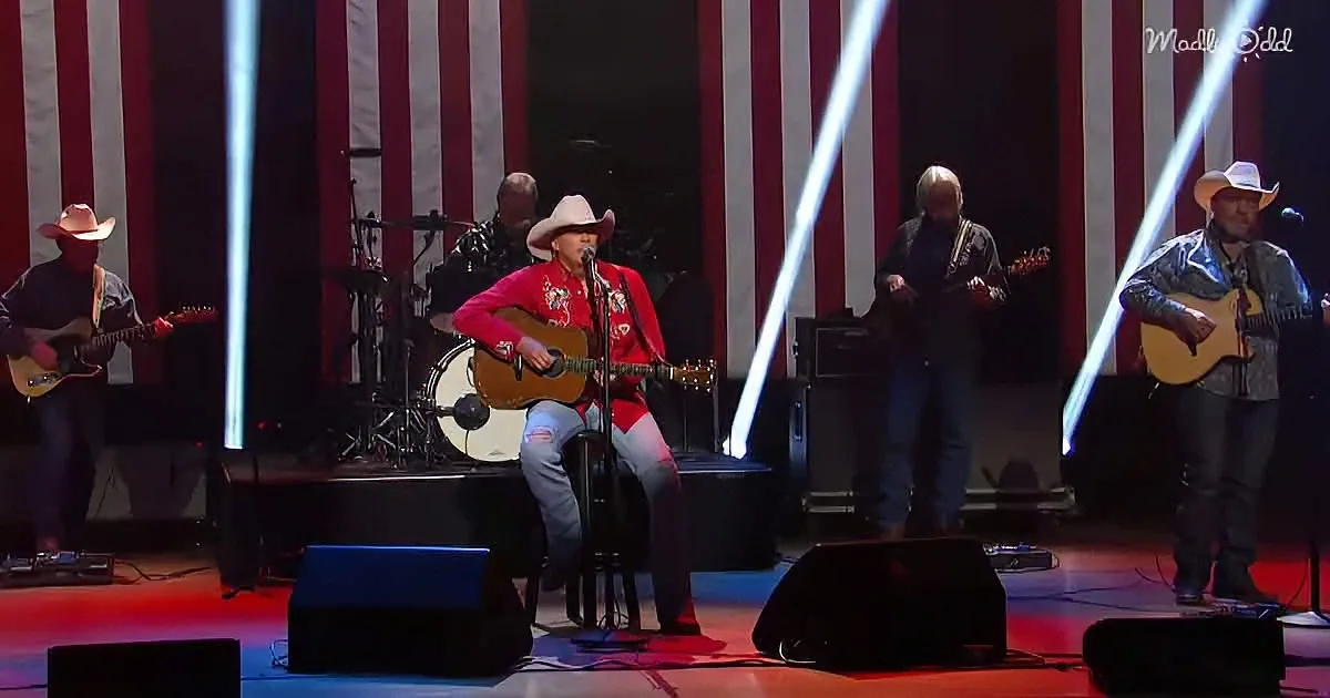 A man in a red shirt and cowboy hat plays guitar on stage with a band and American flag backdrop.