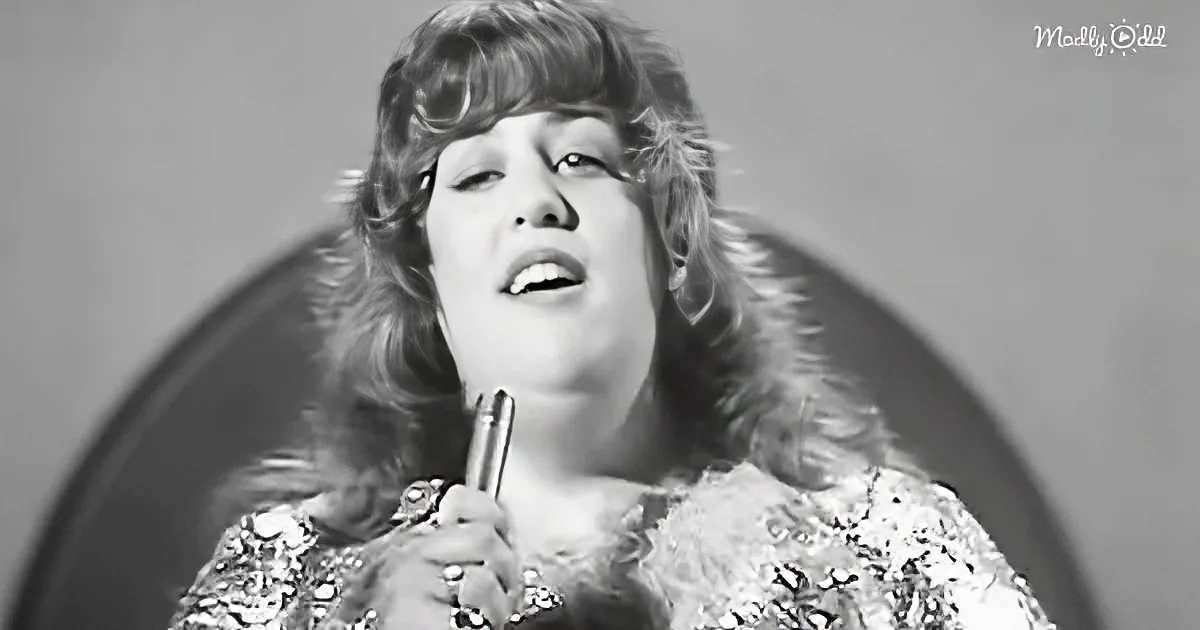 A person with long, wavy hair wearing a sequined top, holding a microphone, singing.