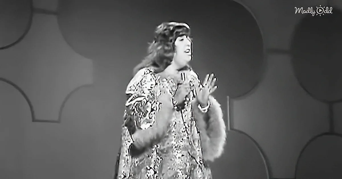 A woman in a fur-trimmed, patterned dress sings on stage with a microphone.