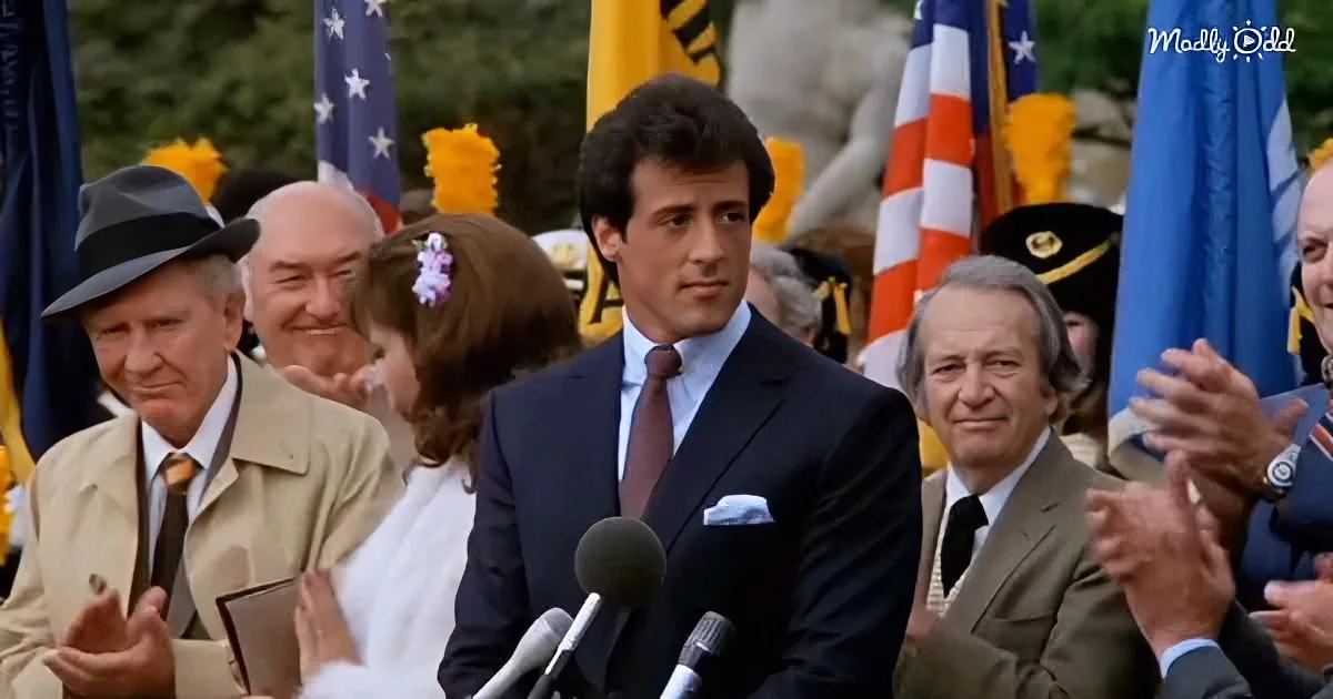 A man in a suit stands at a podium with microphones, surrounded by clapping people and American flags.