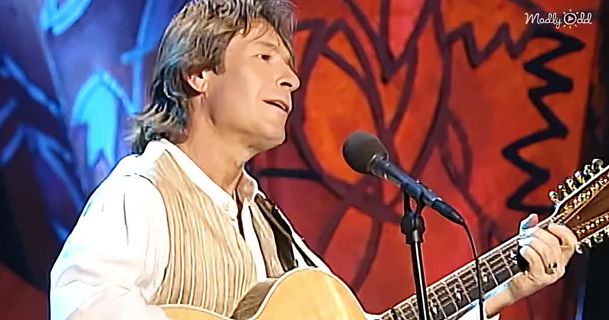 A man plays an acoustic guitar on stage, wearing a white shirt and vest, with a microphone in front of him.