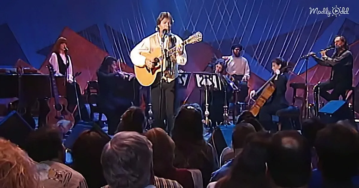 A man plays guitar on stage with a band and audience in front.