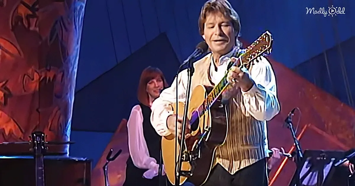 A man in a white shirt and brown vest plays an acoustic guitar on stage, singing into a microphone.