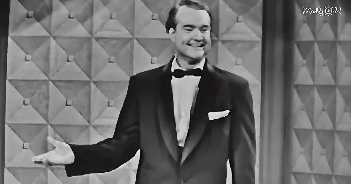 A man in a tuxedo and bow tie smiles, extending his hand, against a patterned backdrop.