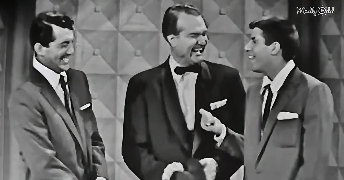A man in a tuxedo gestures while laughing with two other men in suits on a stage.