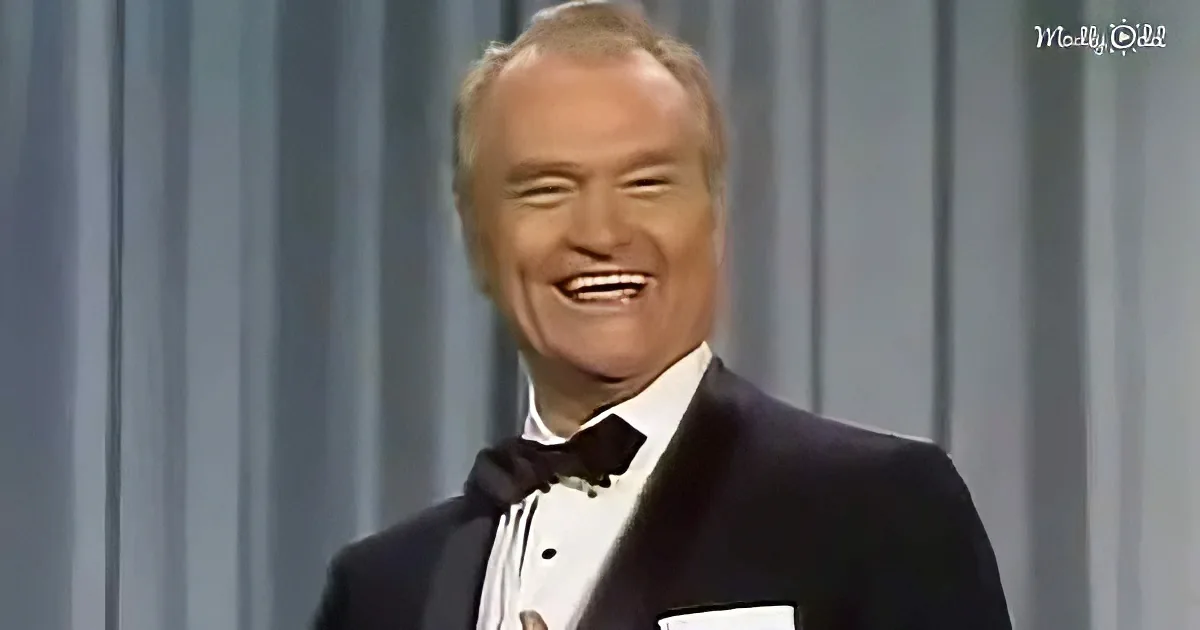 A man in a tuxedo smiles widely, wearing a bow tie, against a gray curtain backdrop.
