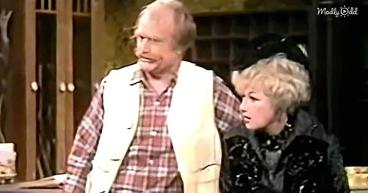 A man in a white vest and plaid shirt stands next to a woman in a black outfit, both looking to the side.