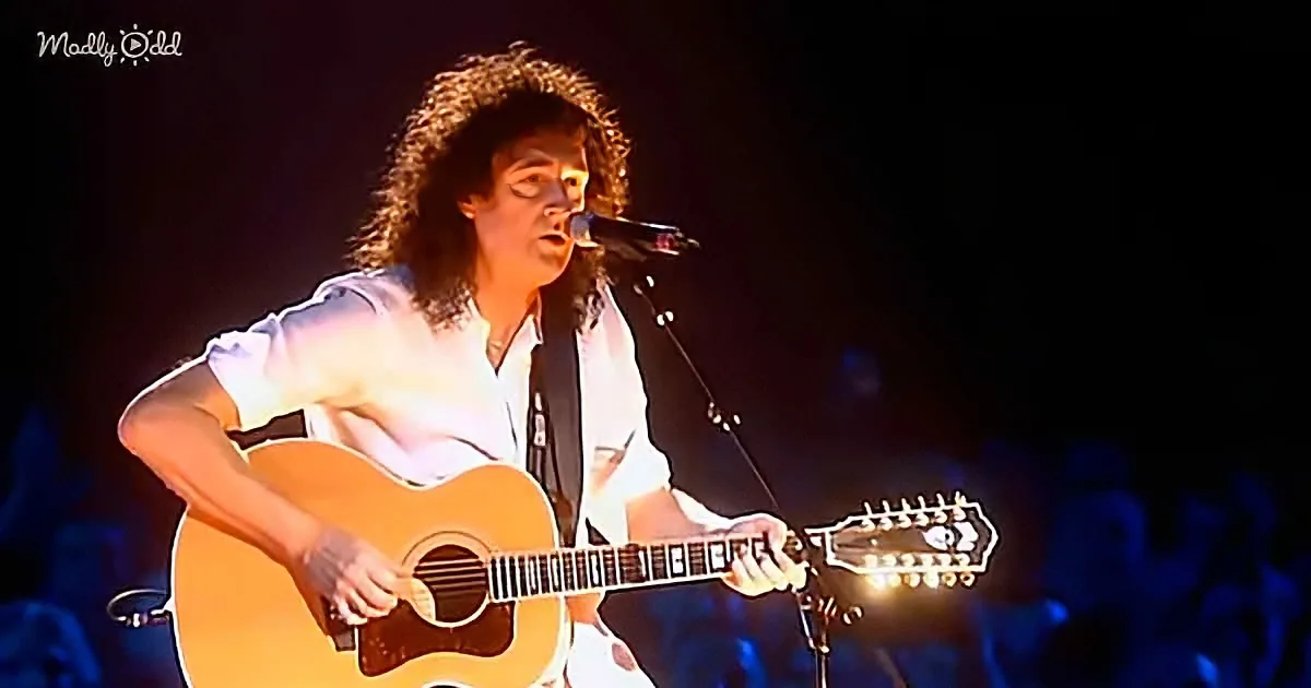 A man with curly hair plays an acoustic guitar on stage, singing into a microphone.