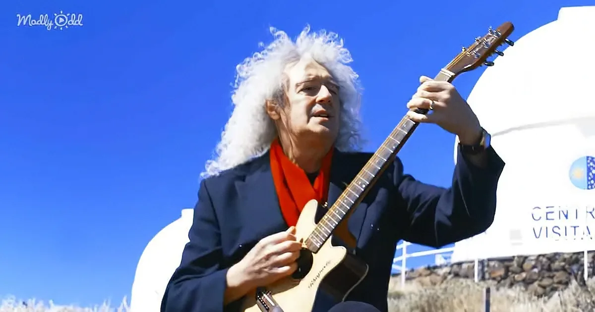 A person with white curly hair plays a guitar outdoors, wearing a black suit and red scarf.