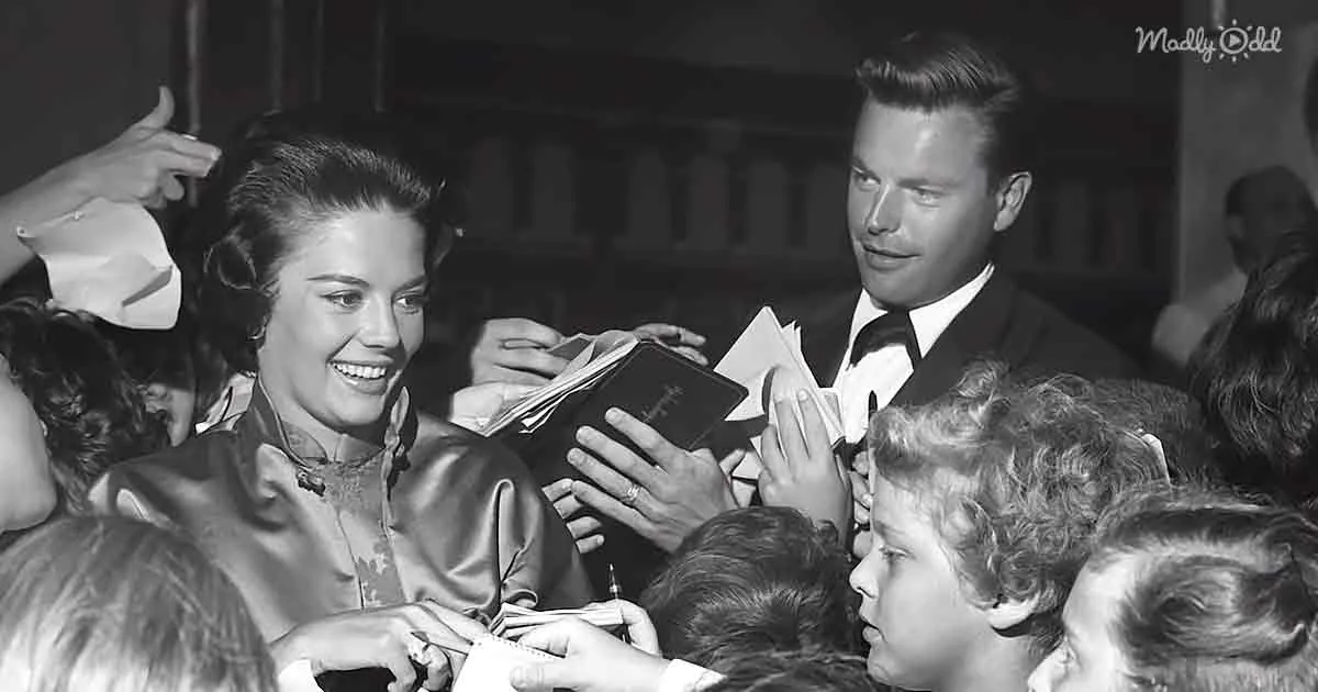 A smiling woman in a satin dress is surrounded by fans, signing autographs and interacting with them.