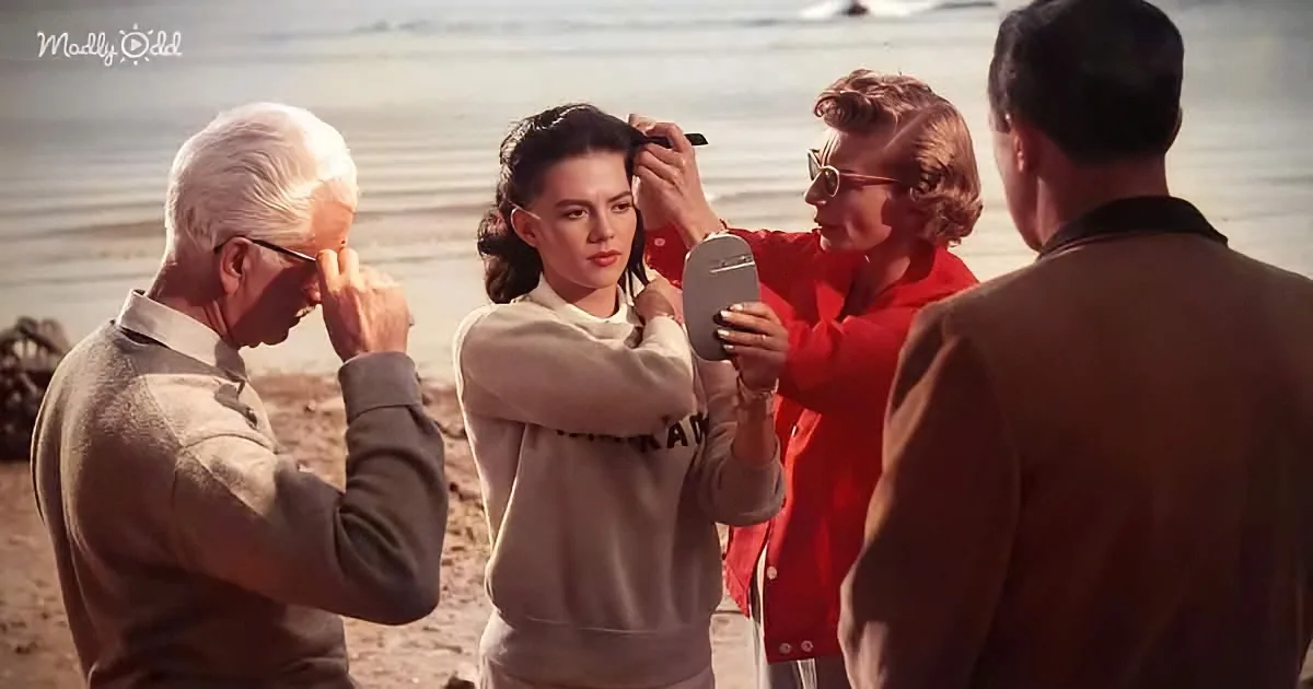 A woman in a beige sweatshirt is getting her makeup done by another woman on a beach.