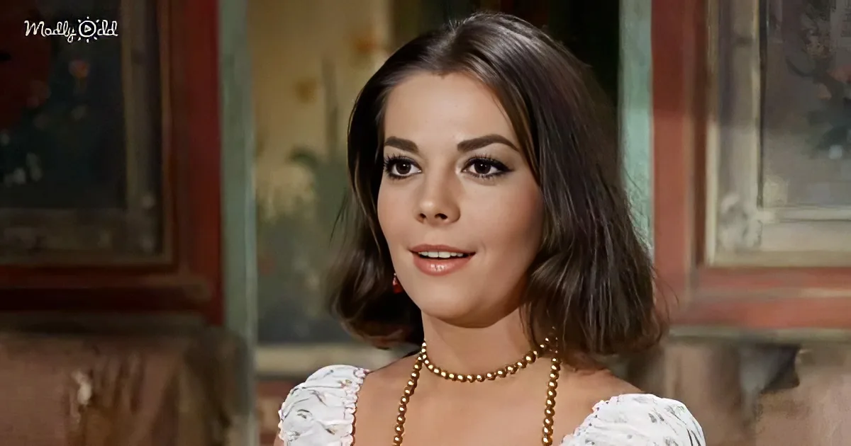 A woman with shoulder-length dark hair, wearing a white dress and a pearl necklace, stands in a room with a painting in...