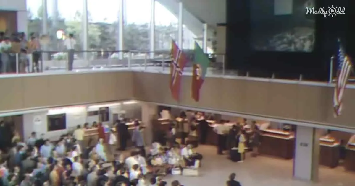 A crowded airport terminal with people waiting, flags hanging, and a large screen in the background.