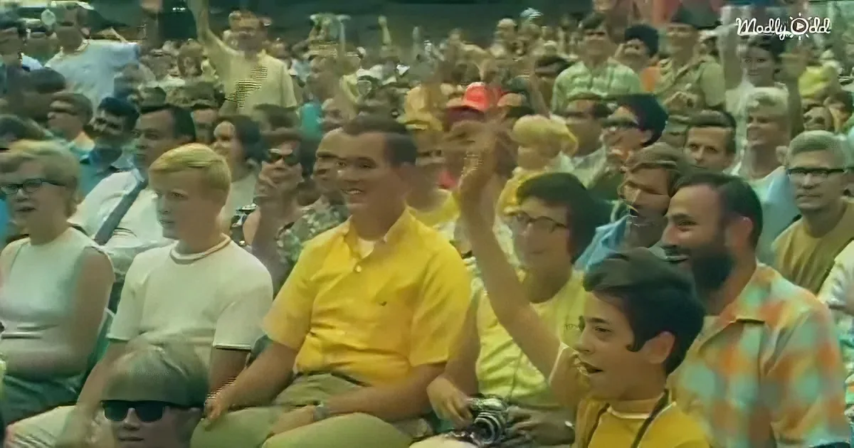 A man in a yellow shirt sits among a crowd, raising his hand, surrounded by people in various attire.