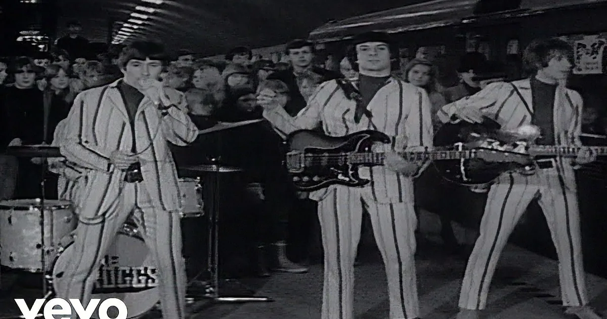 A band in striped suits performs on stage, with a drummer, guitarist, and singer, while a crowd watches.