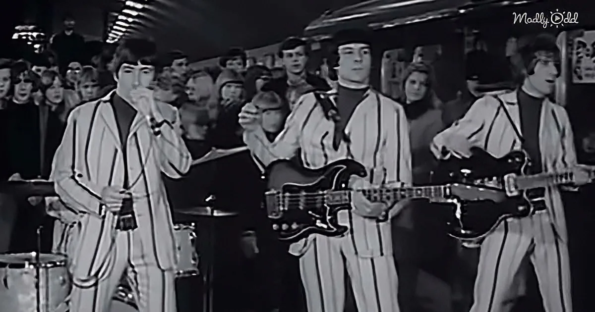 A band in striped suits performs on stage, with a crowd watching and a train visible in the background.