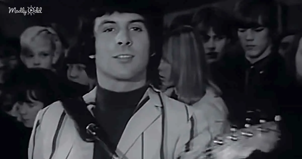 A man in a white jacket and black shirt stands in front of a crowd, holding a guitar.