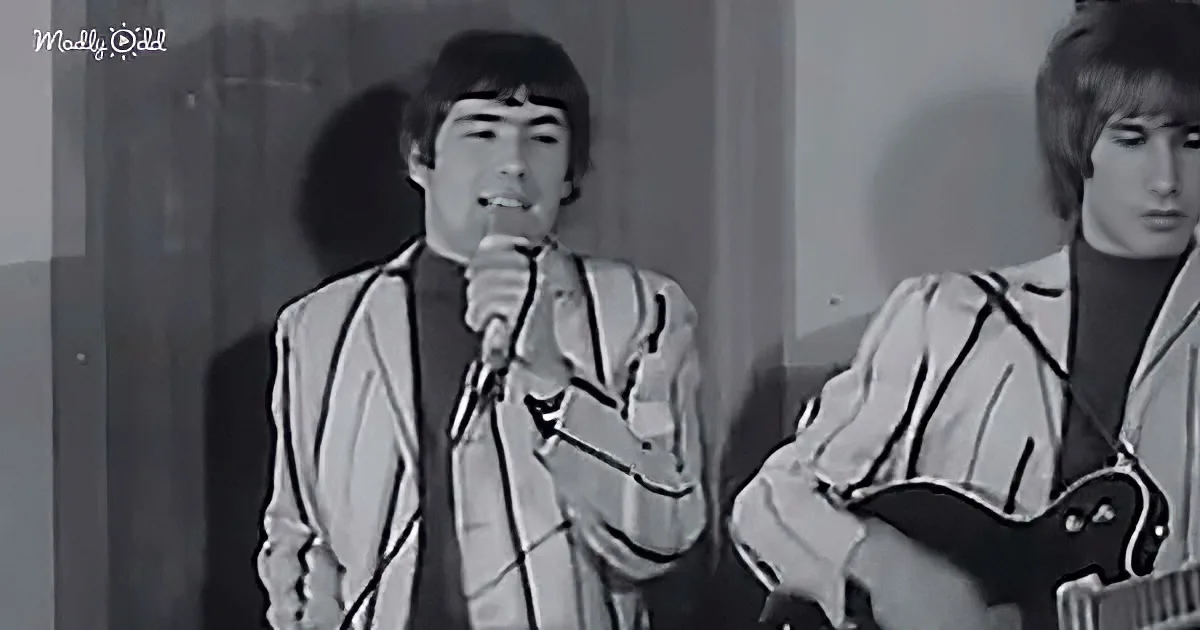A man in a white suit sings into a microphone, accompanied by another man playing a guitar.