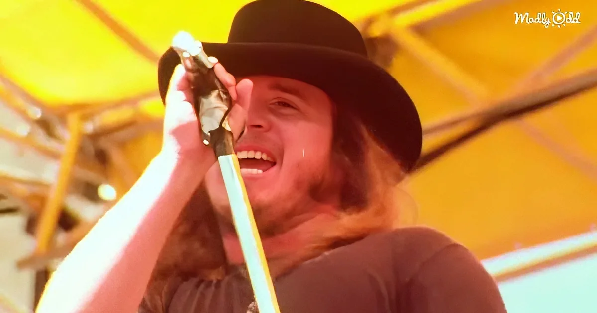 A man with long hair and a hat smiles while holding a microphone under a yellow tent.