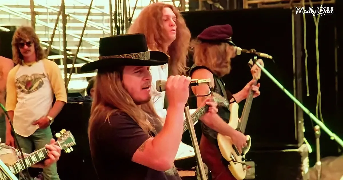 A man with long hair and a hat sings into a microphone while playing guitar on stage.
