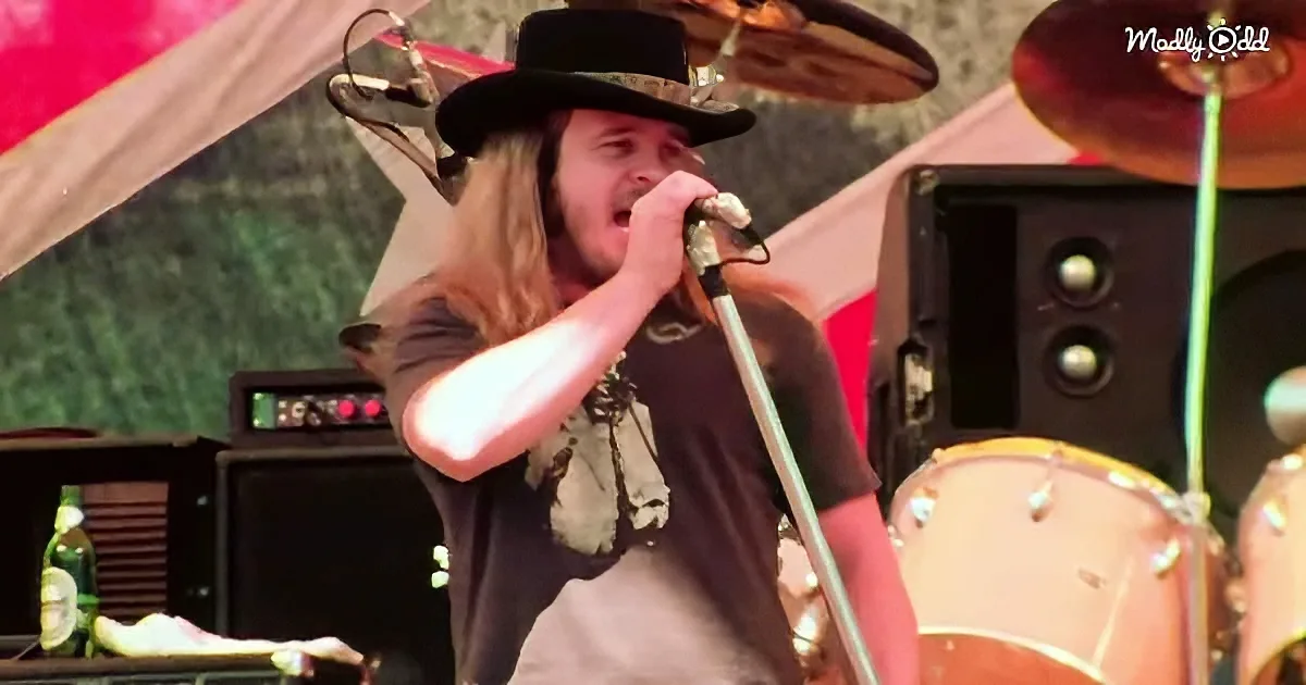 A man with long hair and a hat sings into a microphone on stage, surrounded by musical equipment.