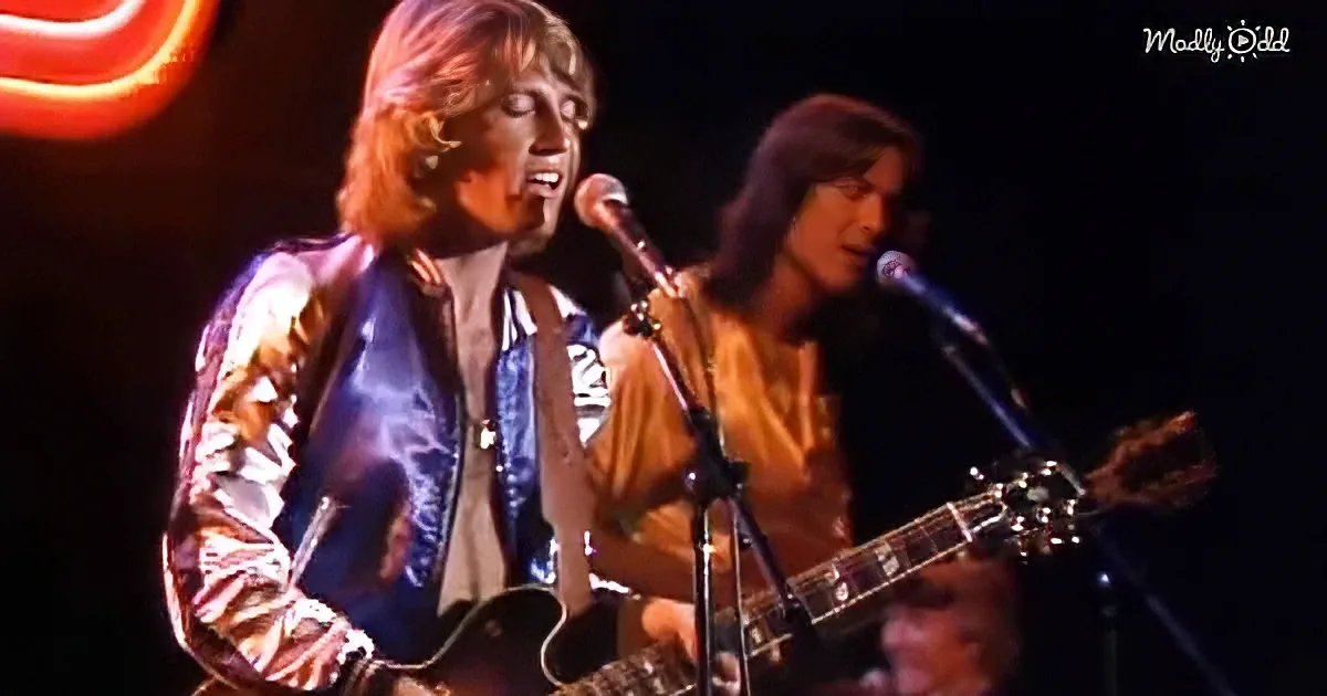 A man in a shiny jacket sings into a microphone while playing guitar on stage.