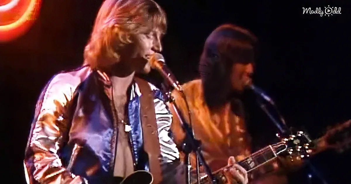 A musician with long hair and a shiny jacket sings into a microphone on stage.