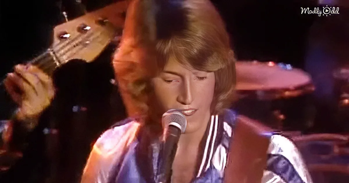 A person with long hair and a striped shirt sings into a microphone, with a guitarist in the background.