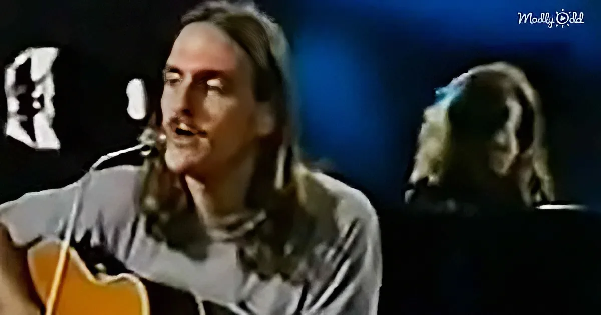 A man with long hair plays guitar and sings into a microphone, with another person in the background.