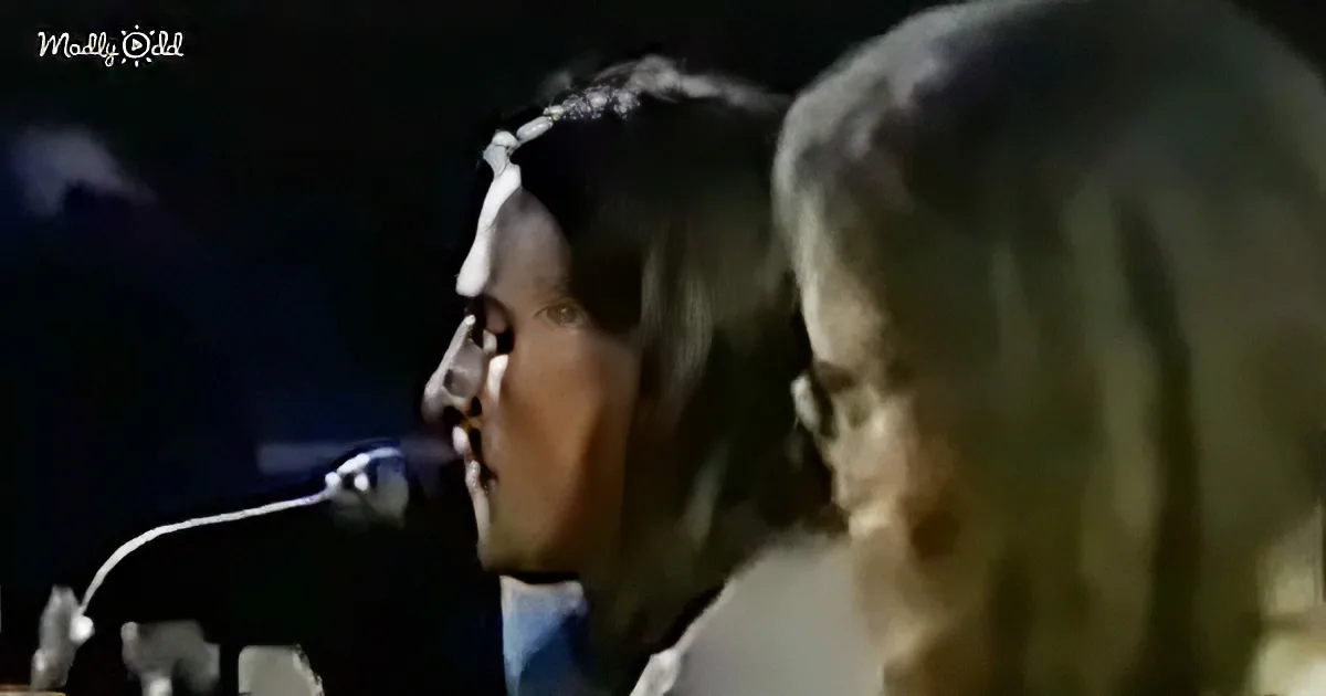 A man singing into a microphone, wearing a dark shirt and tie, with another person partially visible behind him.